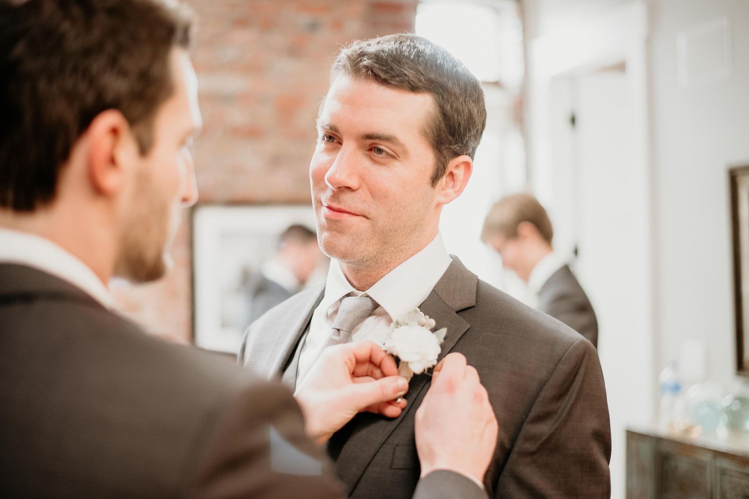 Groomsman pinning a white boutonniere on groom's dark suit lapel before wedding ceremony.