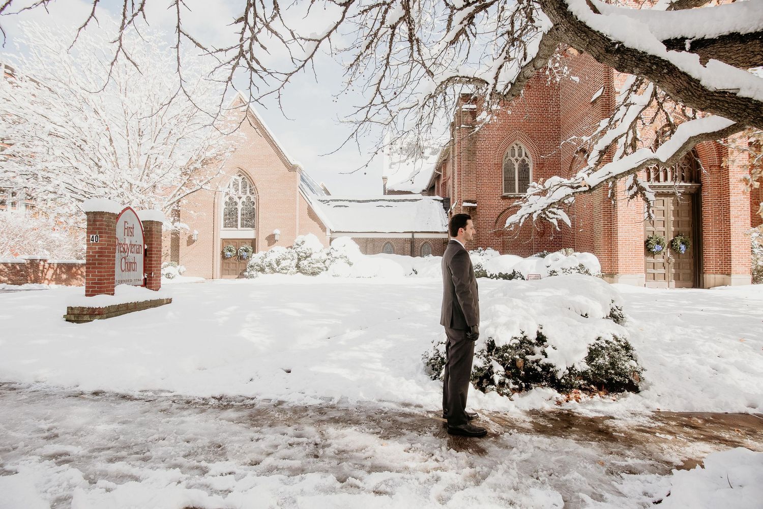 A person stands alone in a snow-covered courtyard beside a red brick church on a winter day.
