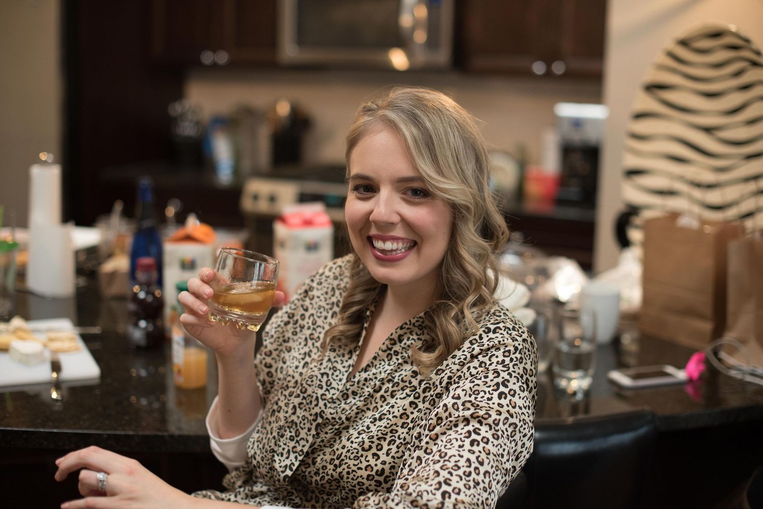 Smiling blonde woman in leopard print top holding a glass of whiskey at a kitchen bar counter.