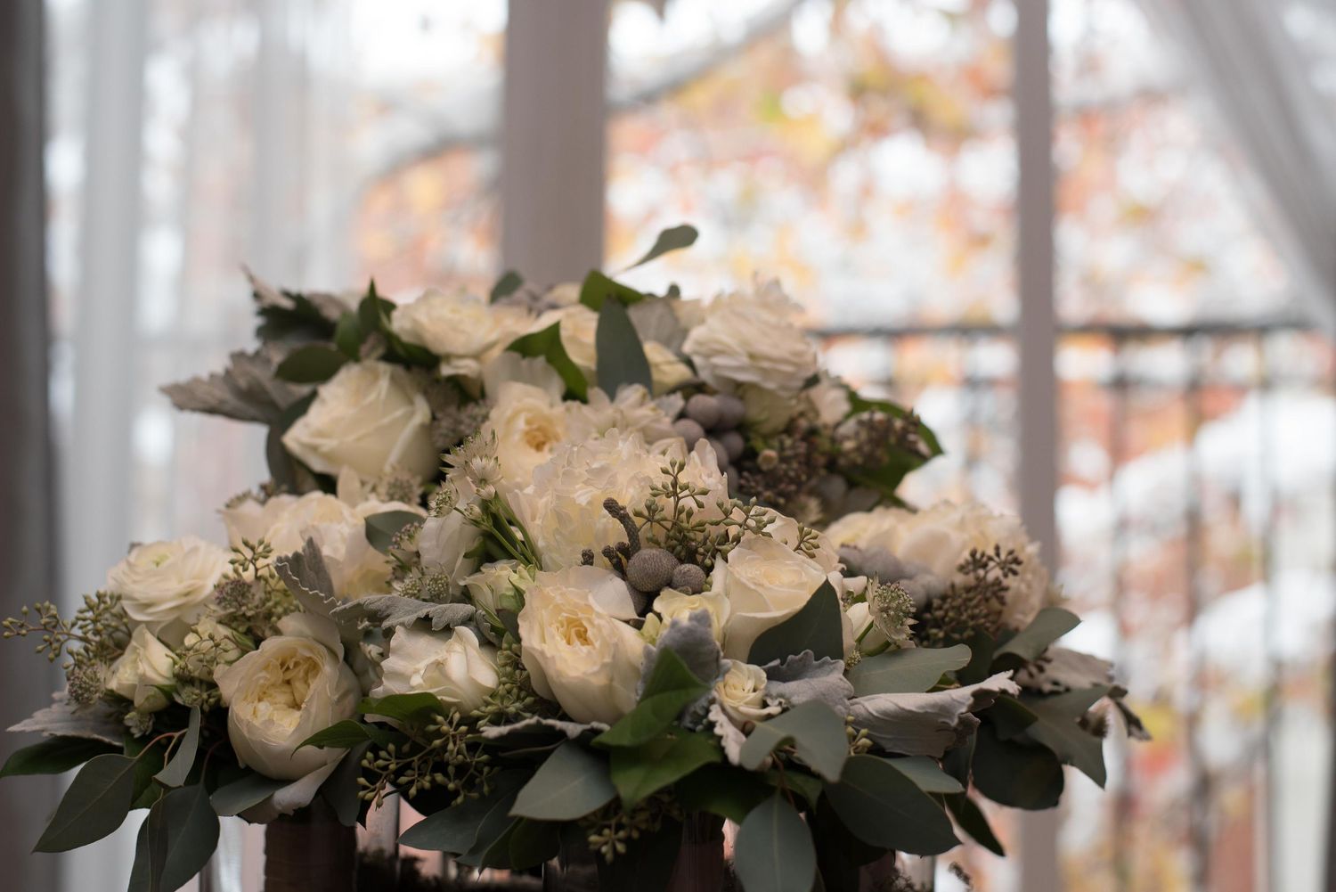 Elegant white rose bouquet with eucalyptus greenery displayed near a window with autumn foliage backdrop.