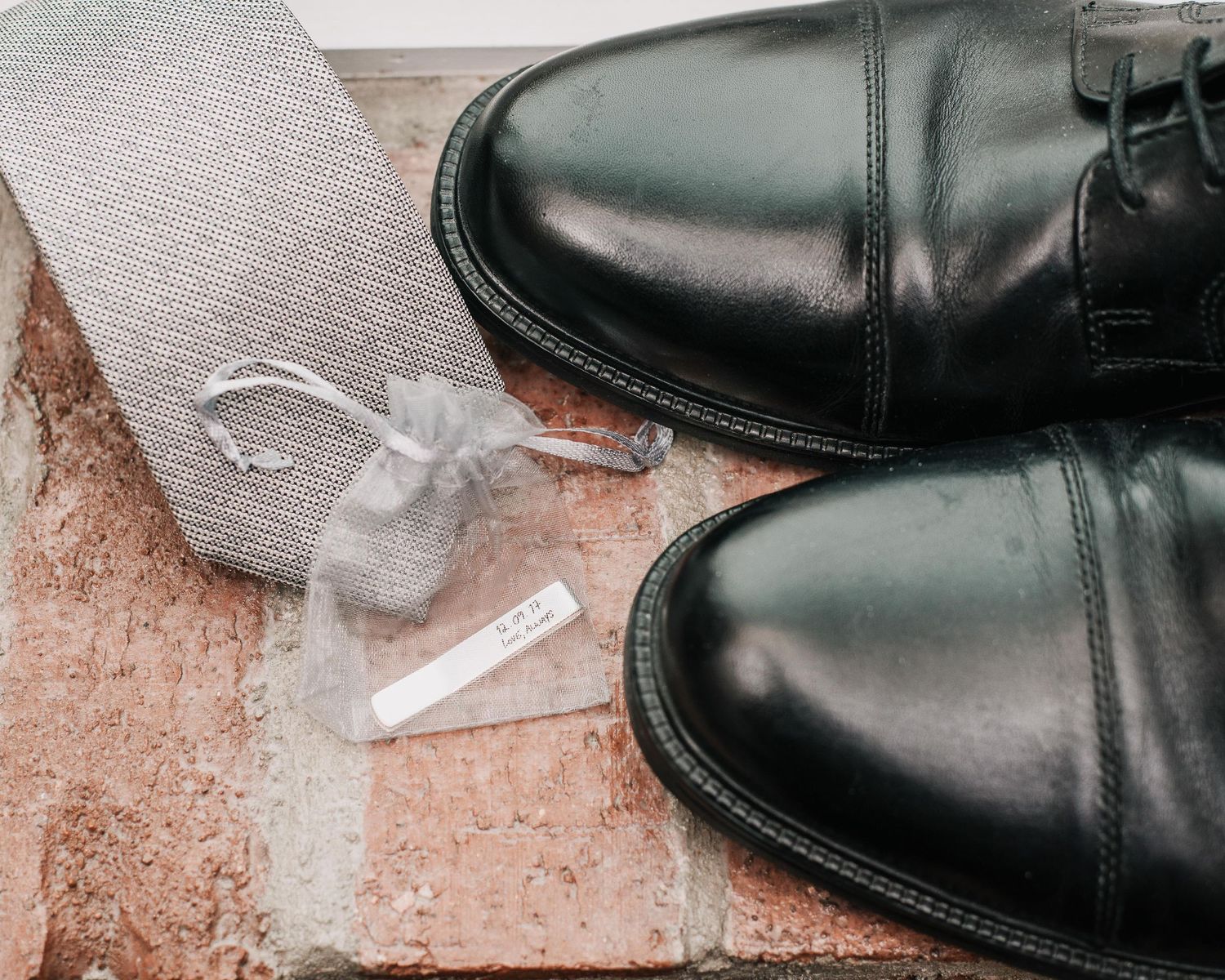 Polished black dress shoes beside a gray tie and small gift bag on a brick surface.