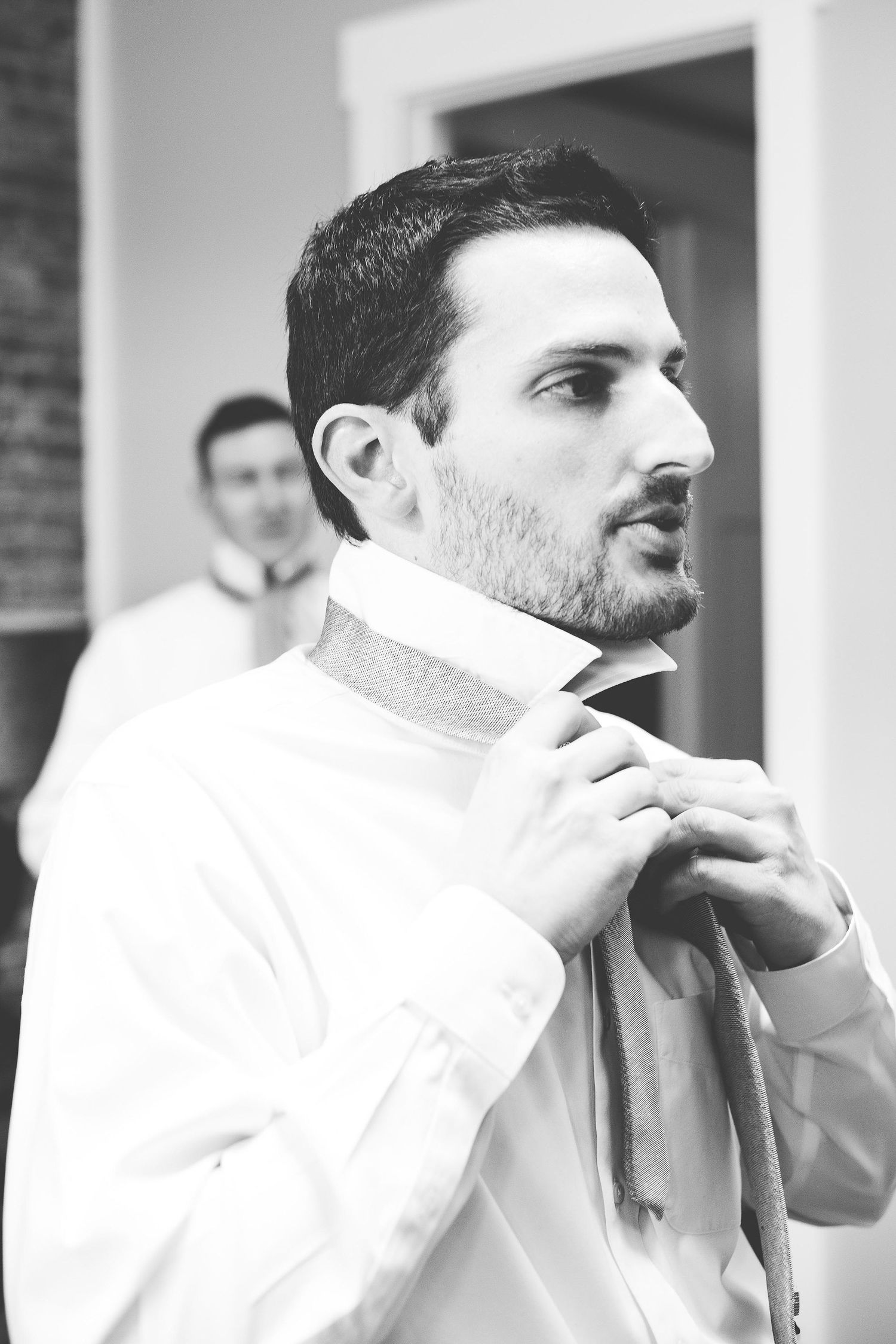 Black and white photo of a groom adjusting his tie while getting ready, with a groomsman visible in the background.