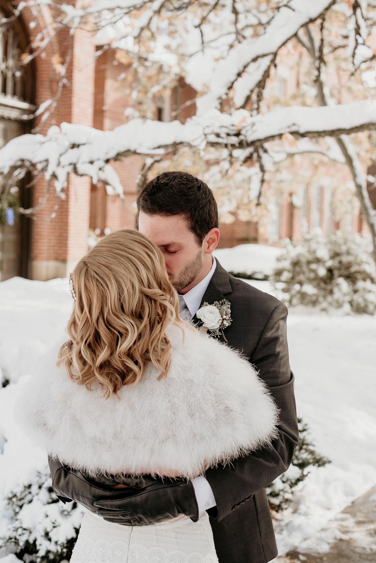 Bride and groom share a kiss outdoors in winter snow, bride wearing white fur wrap shawl, groom in dark suit with boutonniere.