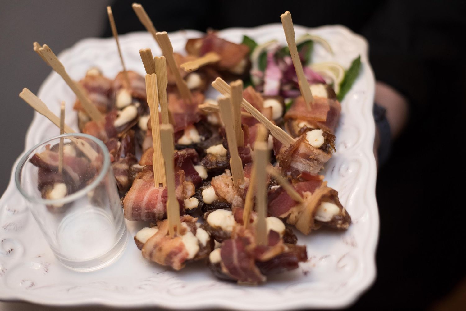 Elegant white platter of bacon-wrapped appetizers with toothpicks and crumbled cheese, served at a catered event.