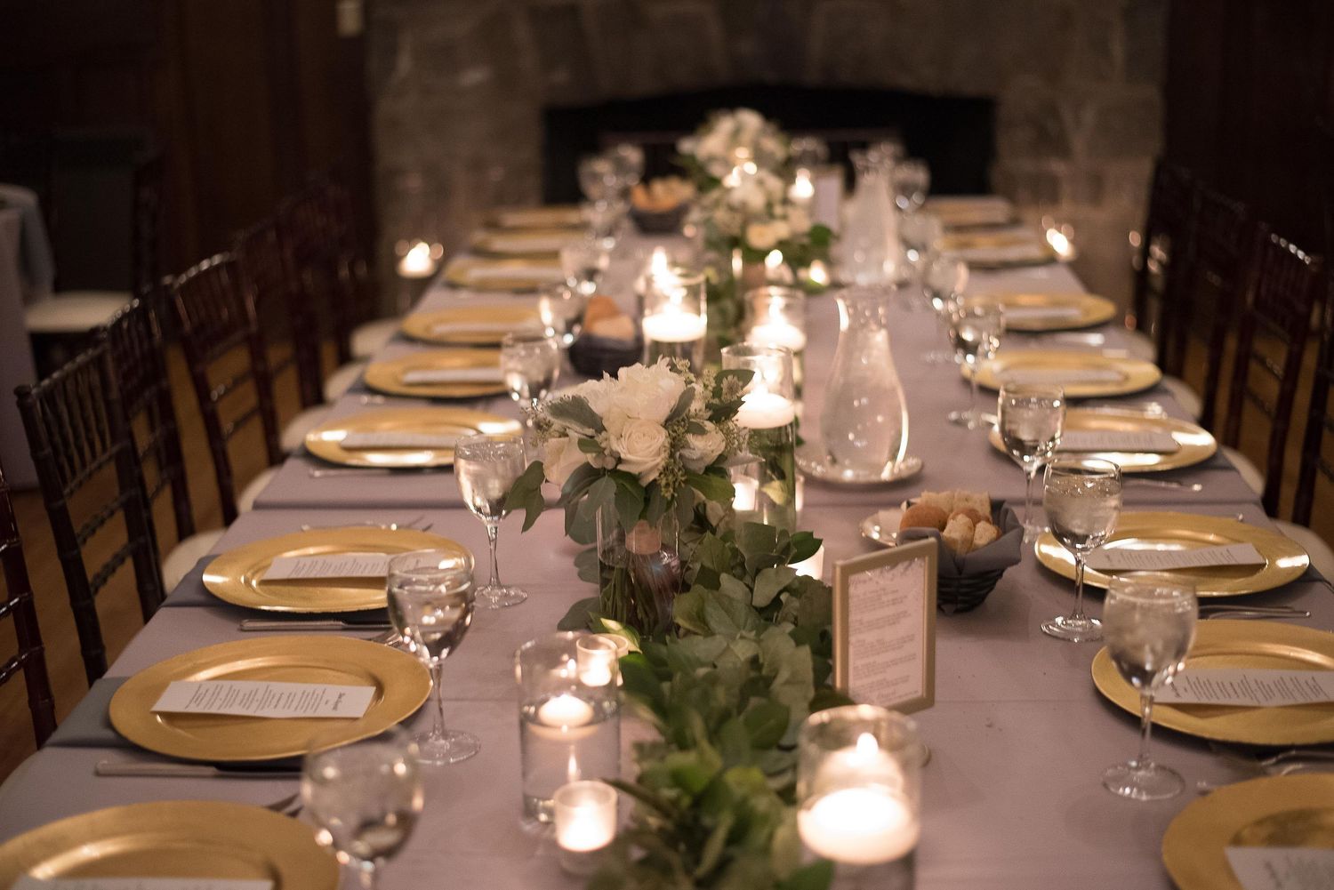 Elegant wedding reception table with gold chargers, candles, and greenery centerpieces in a cozy stone fireplace venue.