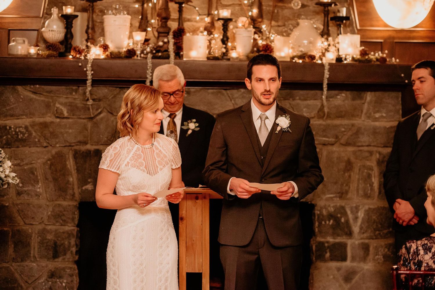 Couple exchanging vows at indoor wedding ceremony beside stone fireplace with candles and floral decor.