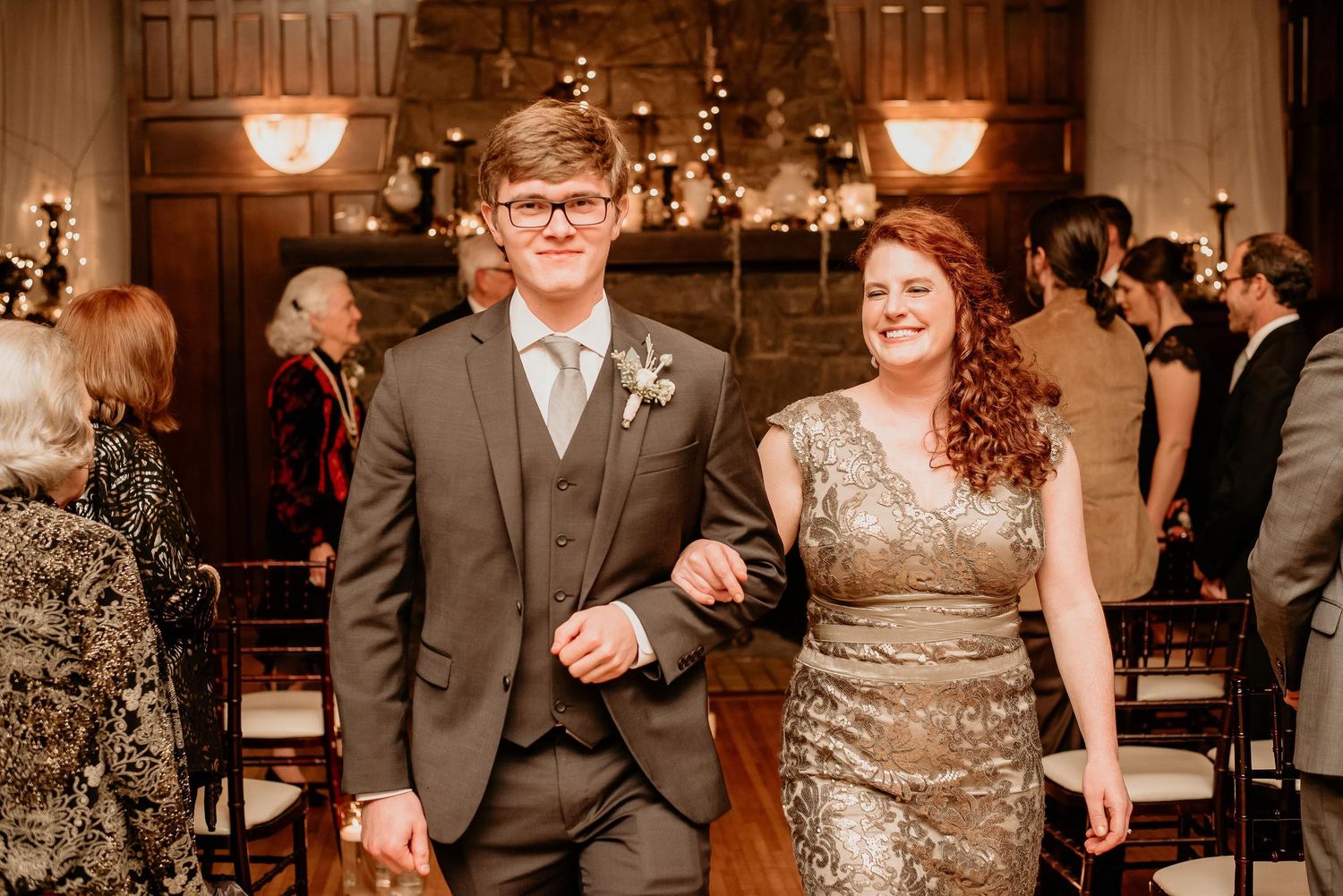 Young couple in formal attire walking down wedding aisle, man in gray suit, woman in gold sequin dress.