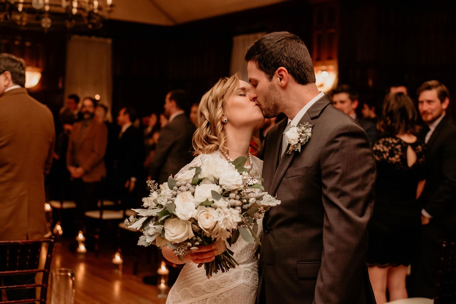 Bride and groom share first kiss at wedding ceremony, bride holding white floral bouquet in elegant indoor venue.