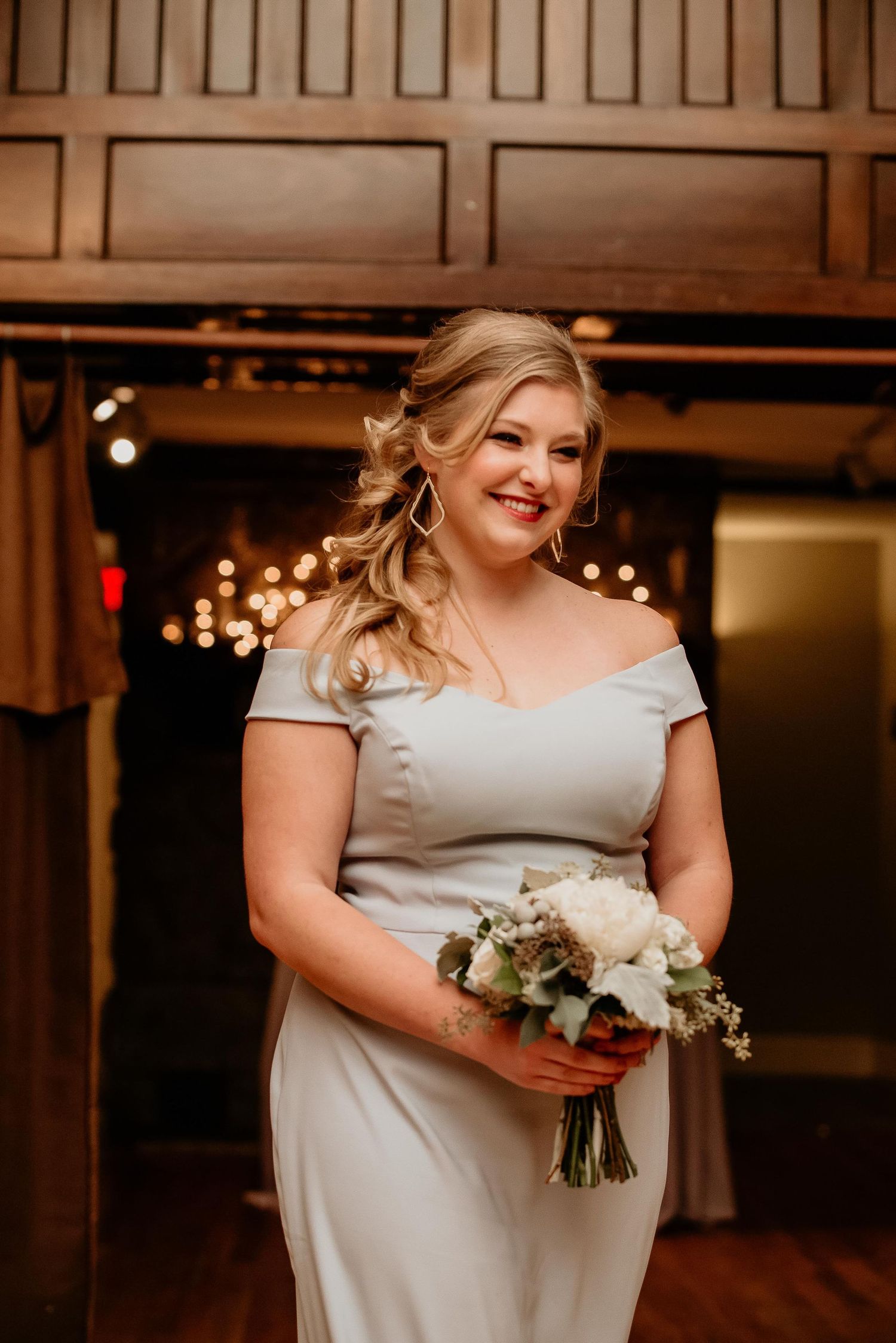 Smiling bridesmaid in off-shoulder silver gown holding white floral bouquet in elegant warmly lit venue.
