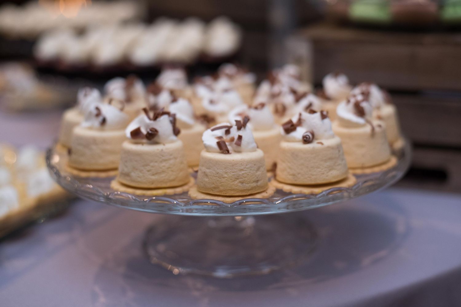 Mini cheesecakes topped with whipped cream and chocolate shavings displayed on a glass cake stand.