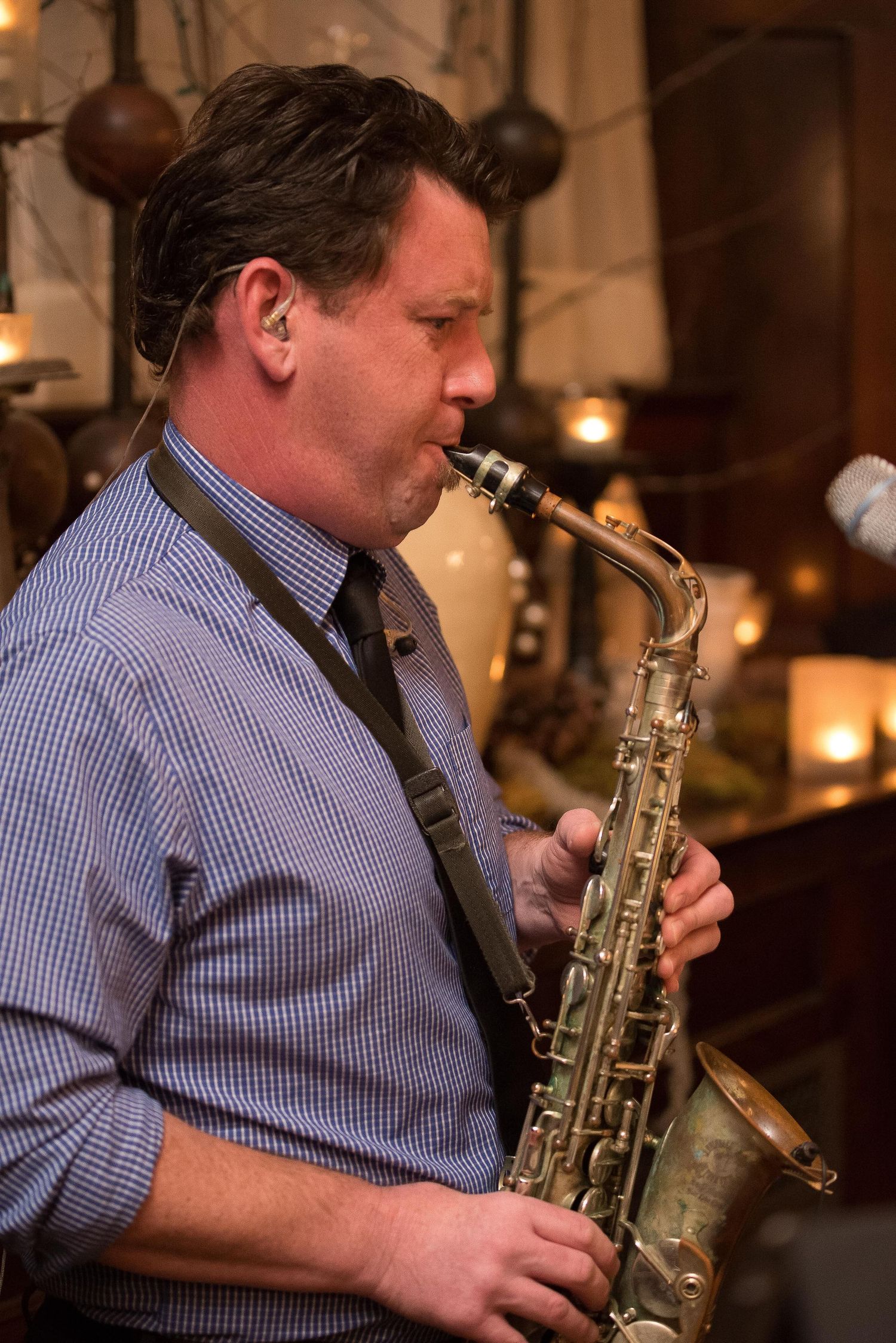 A man in a blue checkered shirt plays a brass saxophone at a candlelit jazz event.