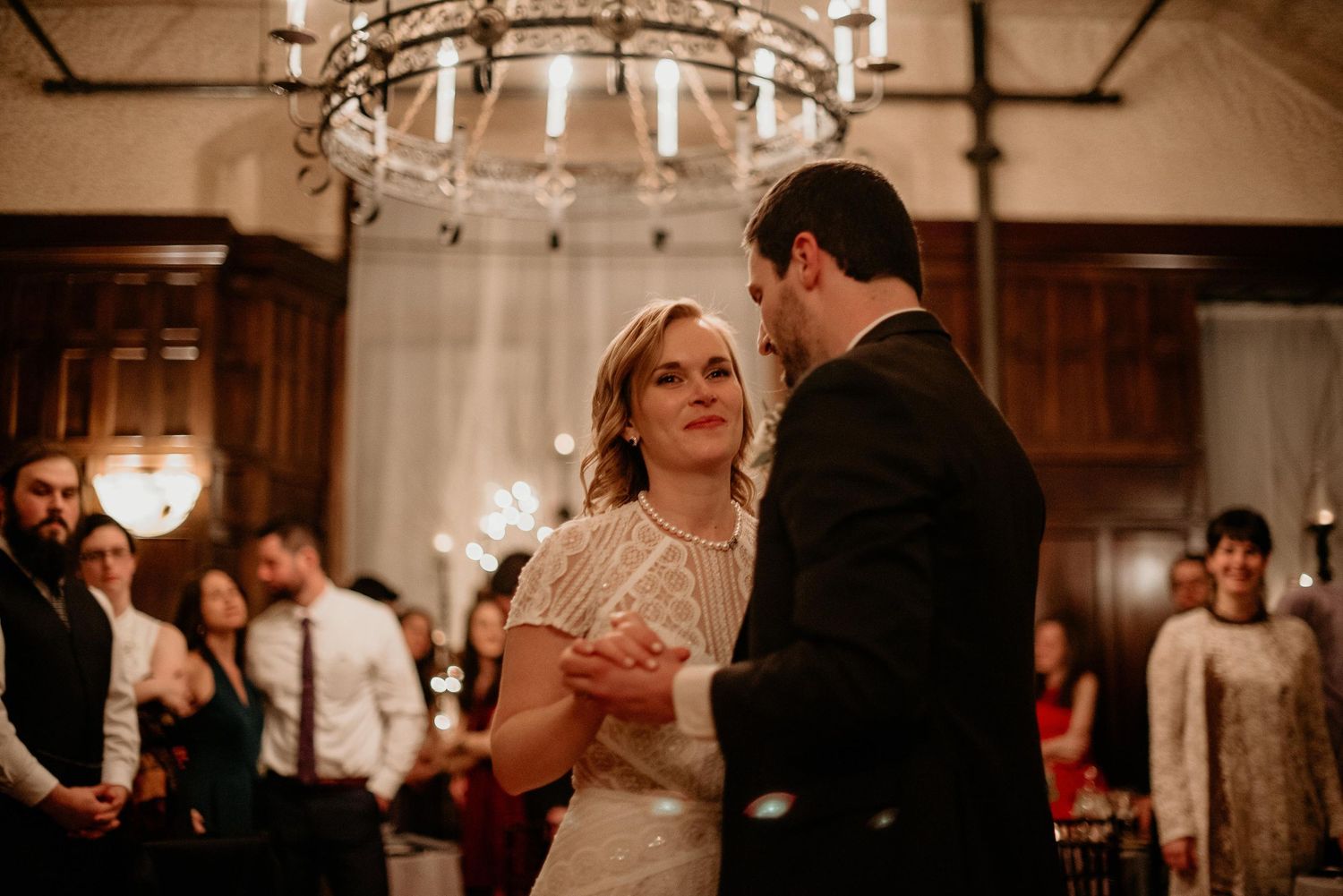Couple shares first dance under chandelier at elegant wedding reception with guests watching.