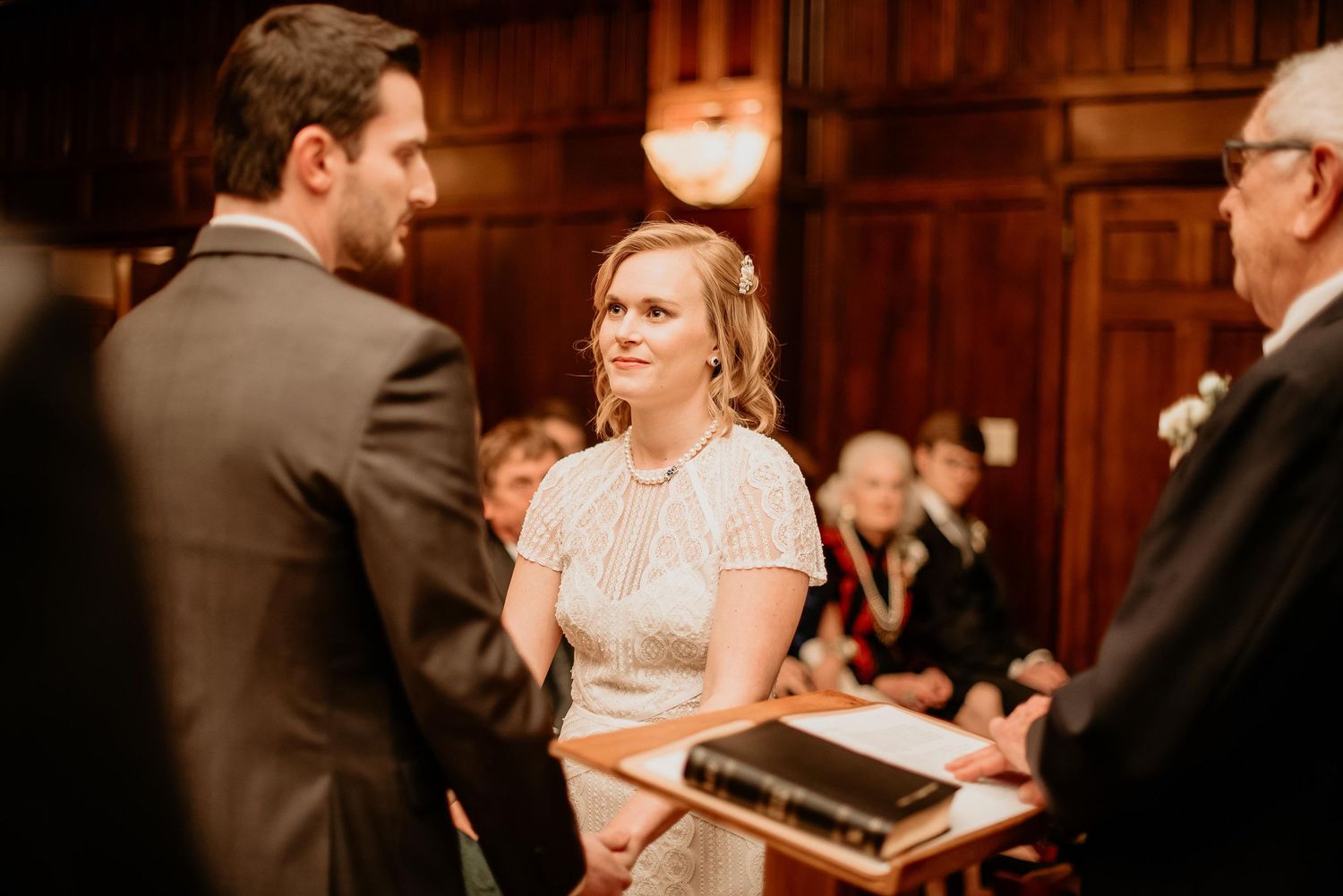 Bride smiling at groom during intimate wedding ceremony at elegant wood-paneled venue with guests seated behind.