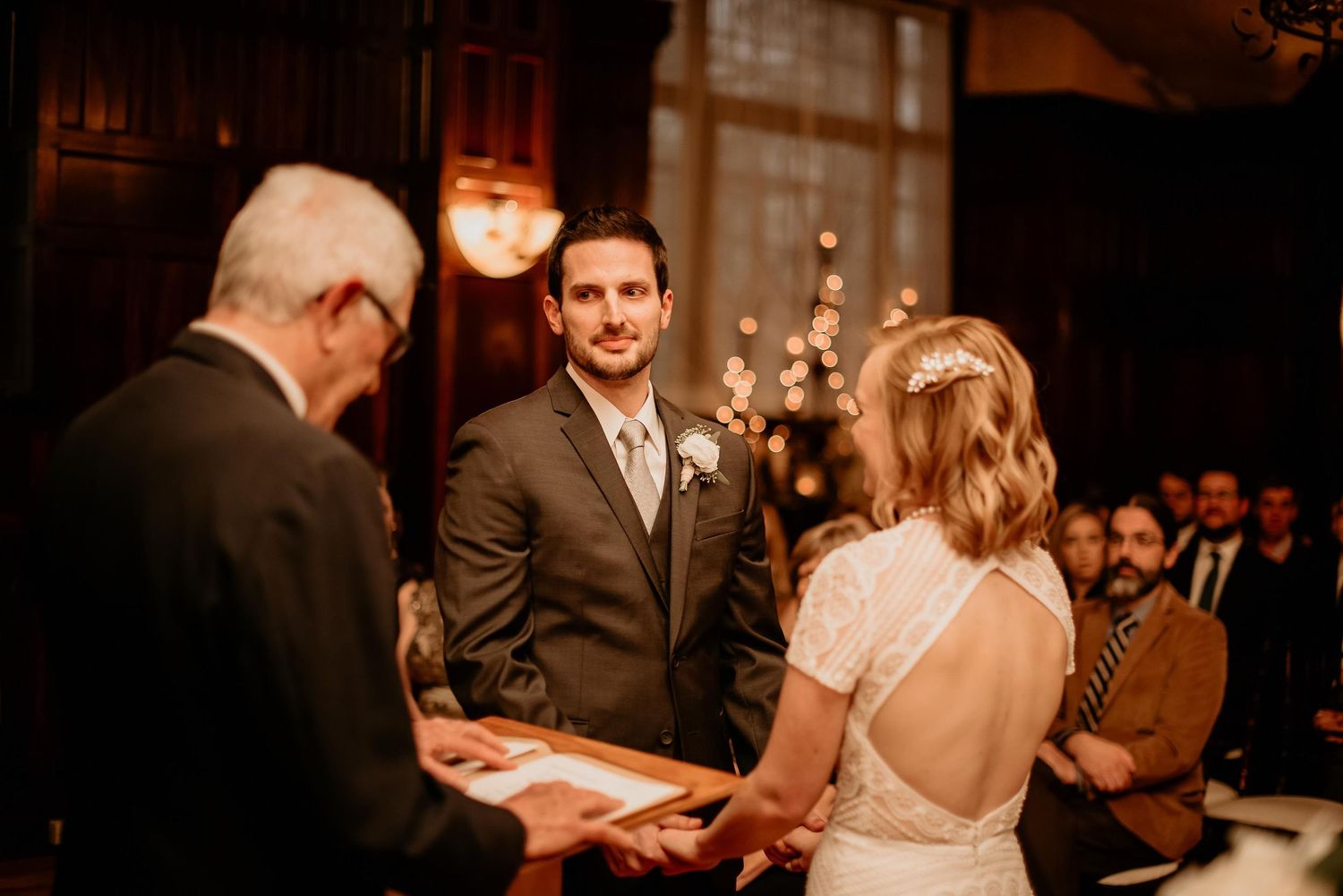 Groom smiling at bride during indoor wedding ceremony with warm bokeh lights in elegant venue.