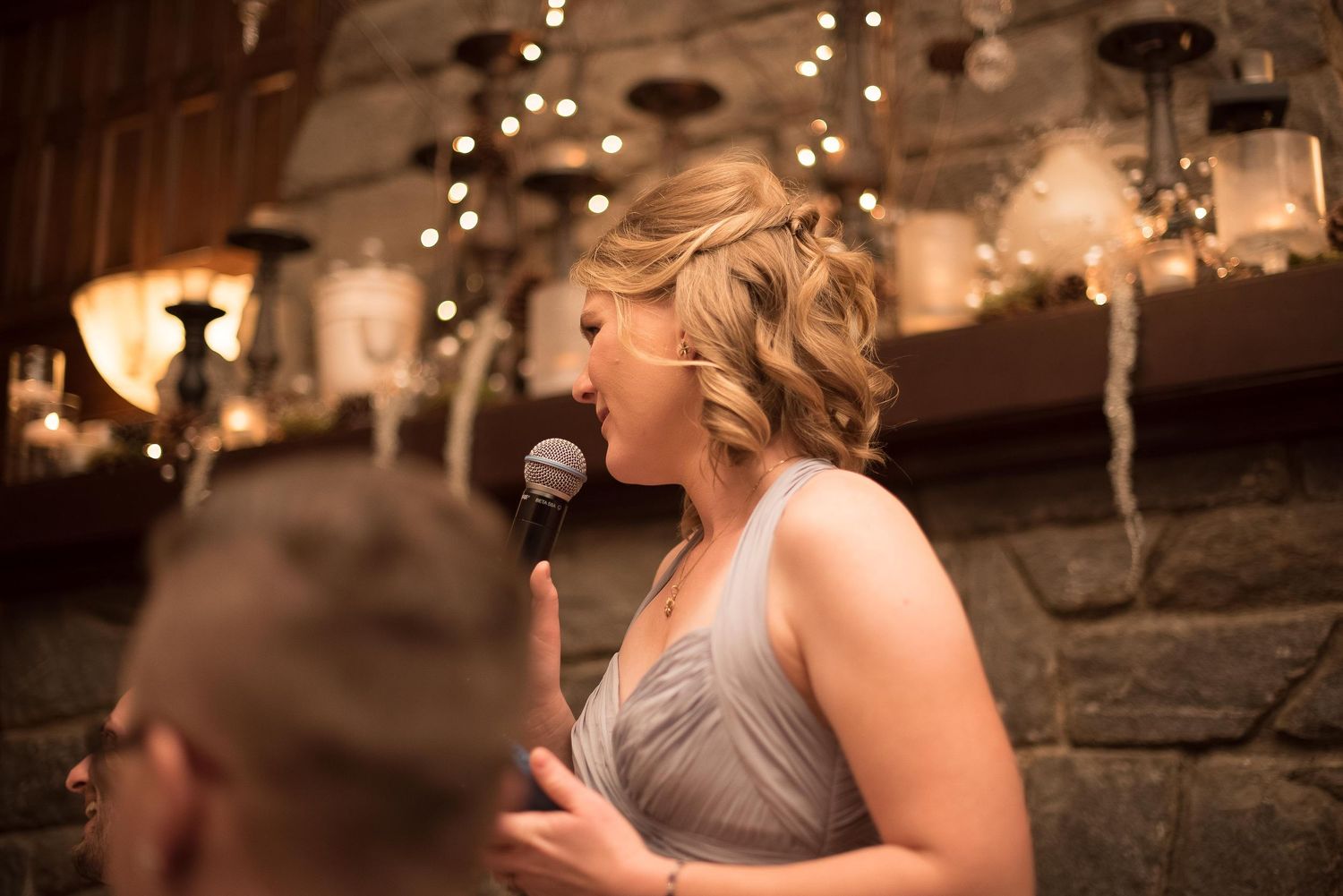 Bridesmaid in gray dress giving a heartfelt speech with microphone at elegant wedding reception with string lights.