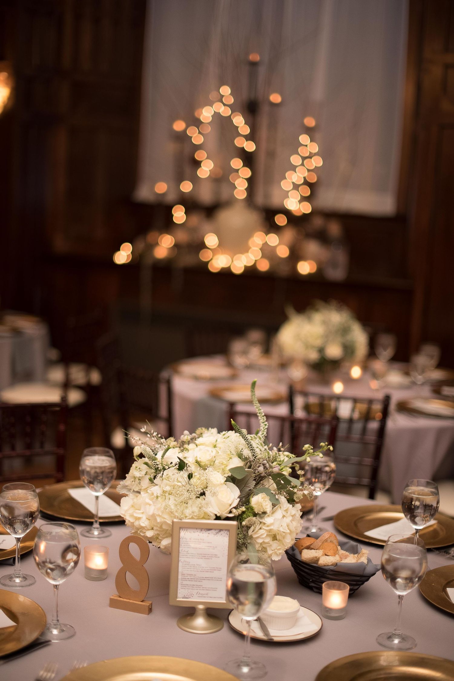 Elegant wedding reception table with white floral centerpiece, gold chargers, candles, and a table number 8 sign.
