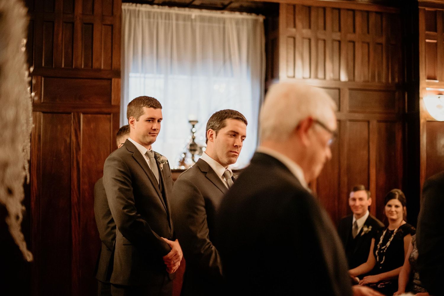Groomsmen in gray suits standing attentively during indoor wedding ceremony.