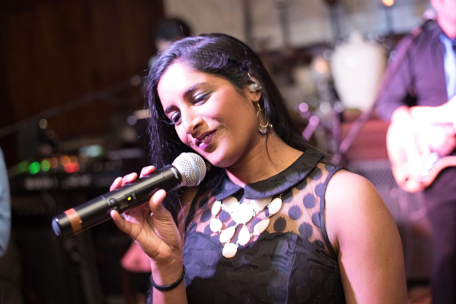 A woman in a black dress sings passionately into a microphone on stage with a band performing behind her.