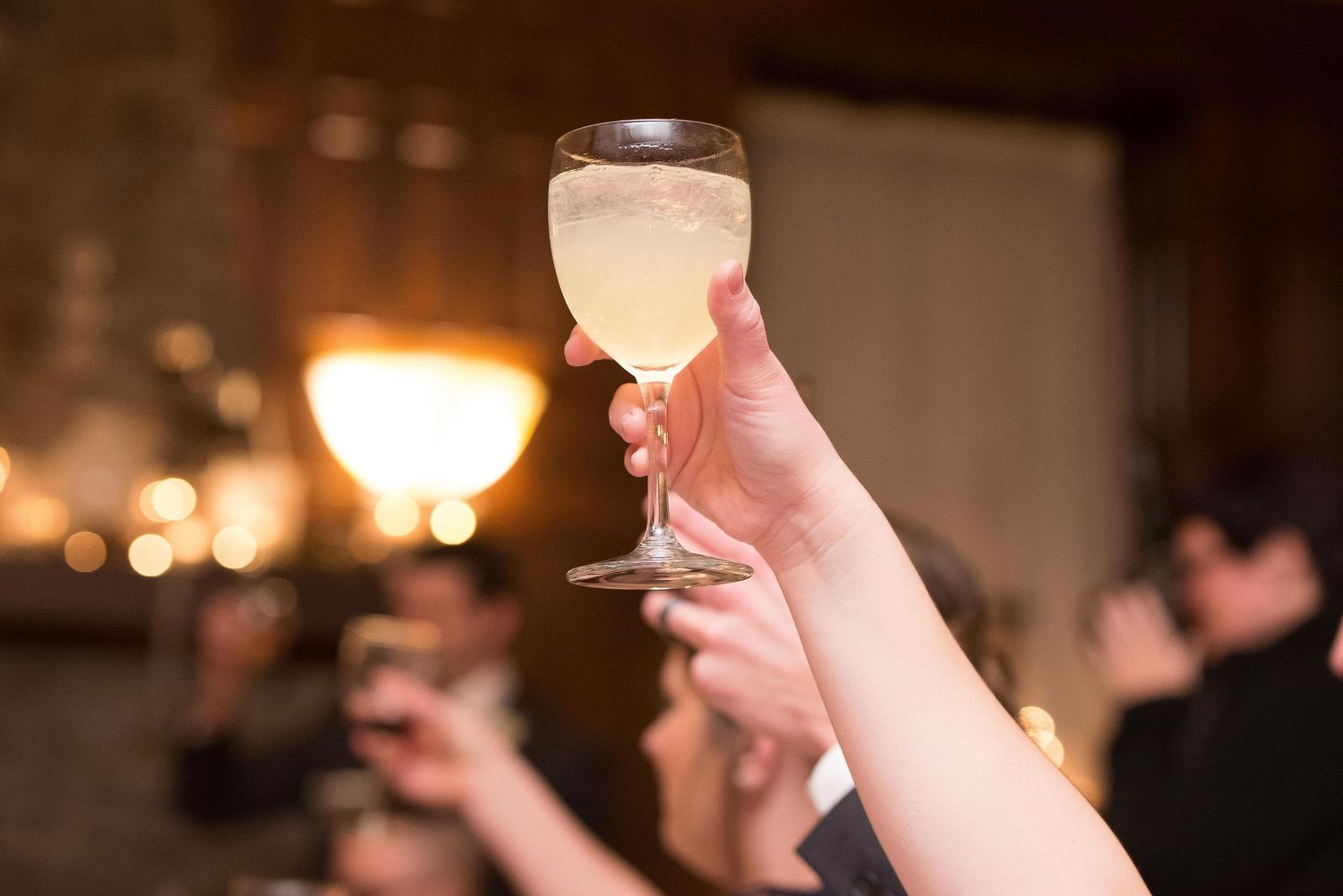 A hand raising a glass of white wine in a toast at an elegant evening celebration or wedding reception.
