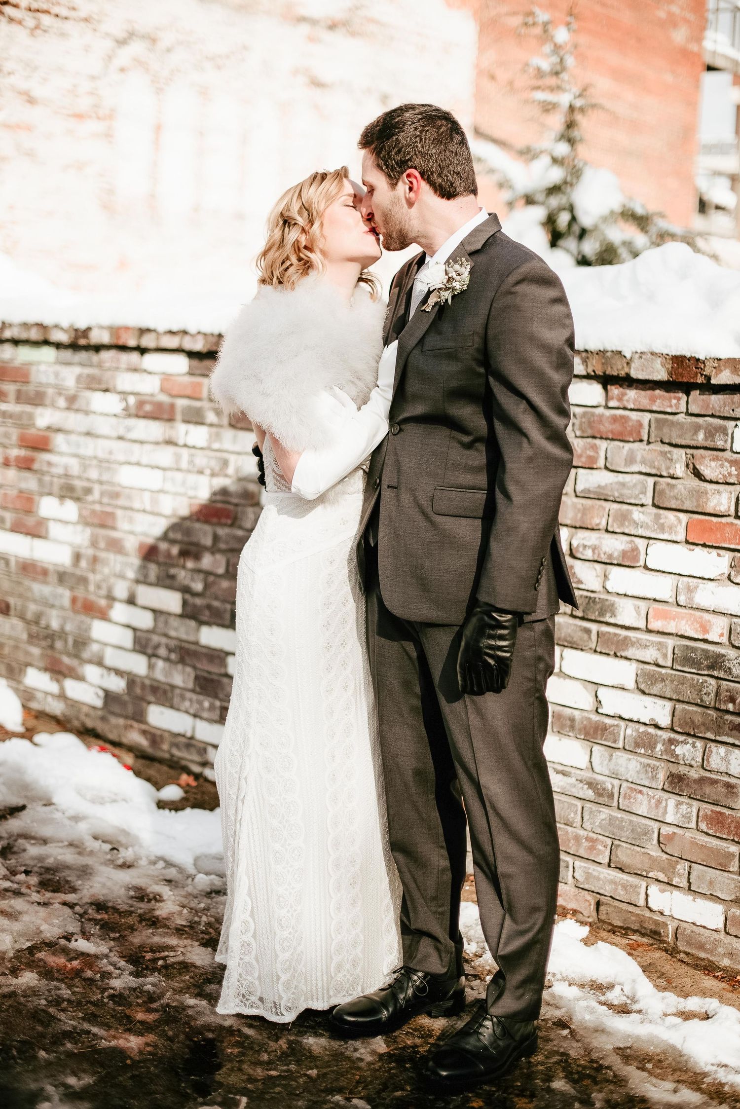 Bride and groom share a kiss in winter wedding attire beside a brick wall dusted with snow.