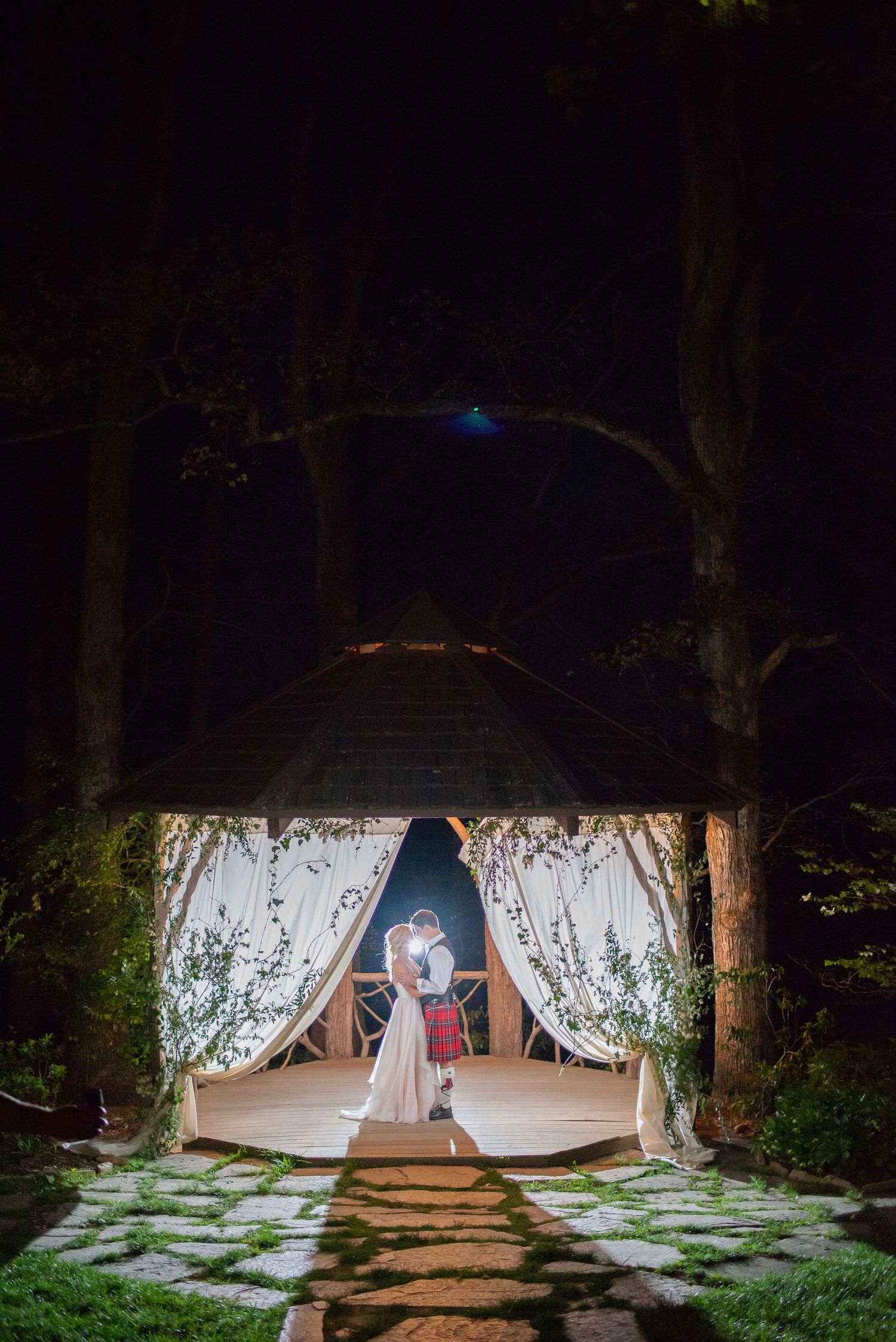 Bride and groom sharing a romantic kiss inside an illuminated gazebo decorated with white drapes at night.