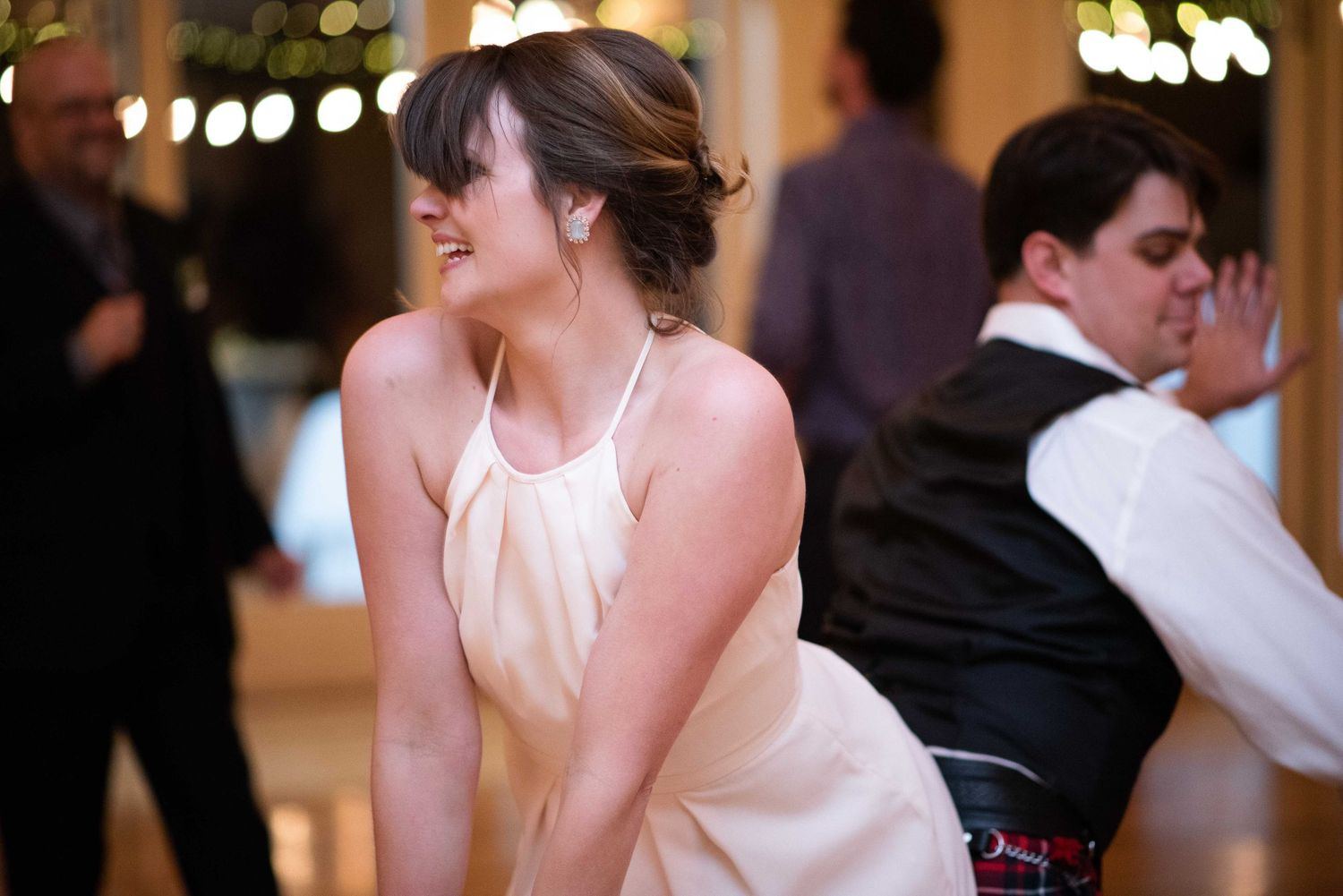 Woman in elegant white halter dress dancing joyfully at a wedding reception with guests in background.