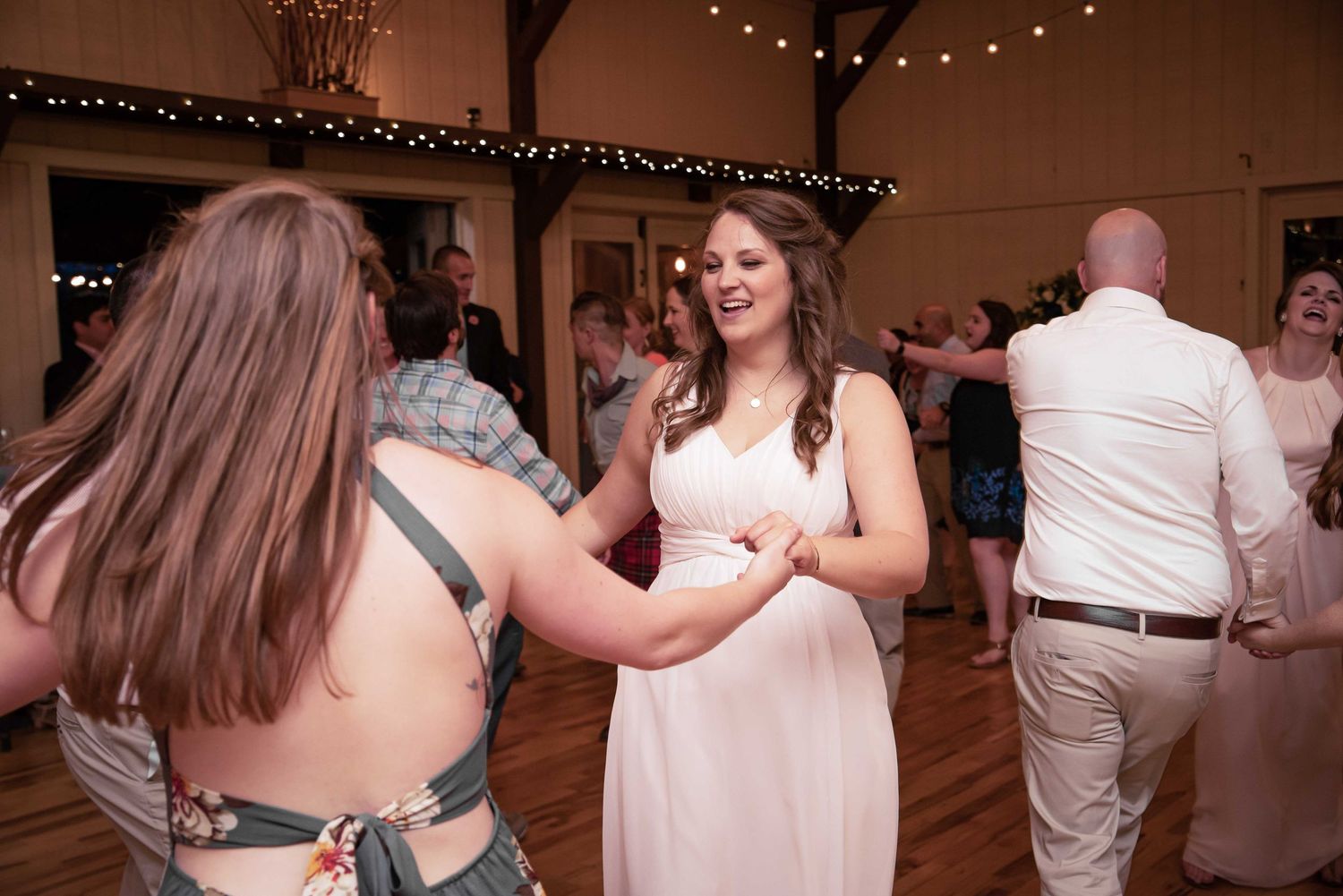 Two women dancing together at a wedding reception in a rustic barn venue with string lights.