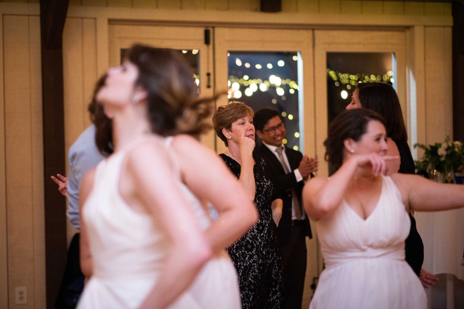 Guests dancing joyfully at a wedding reception inside a warmly lit venue with string lights in the background.