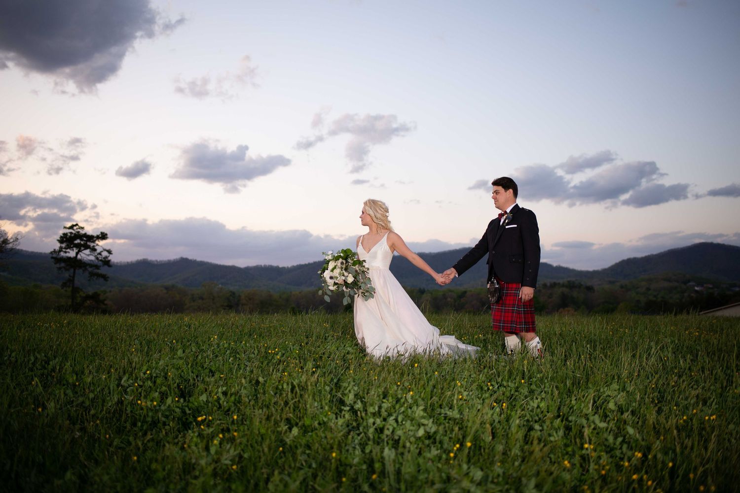 Bride and groom holding hands in a green meadow with mountain views at sunset, groom wearing a red tartan kilt.
