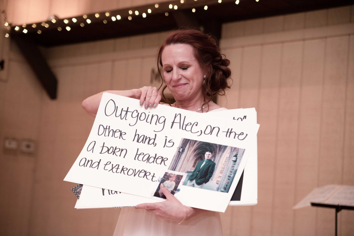 Woman holding a handwritten sign about Alec at a wedding reception with fairy lights in background.