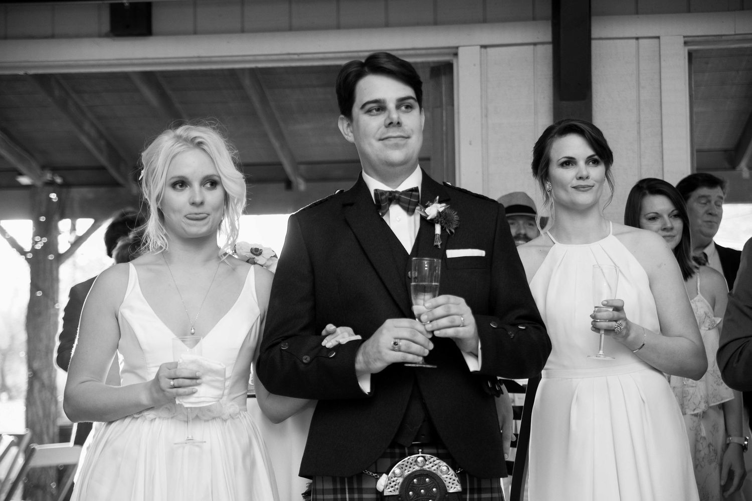 Black and white wedding photo of groom in kilt flanked by two bridesmaids in white dresses holding drinks.