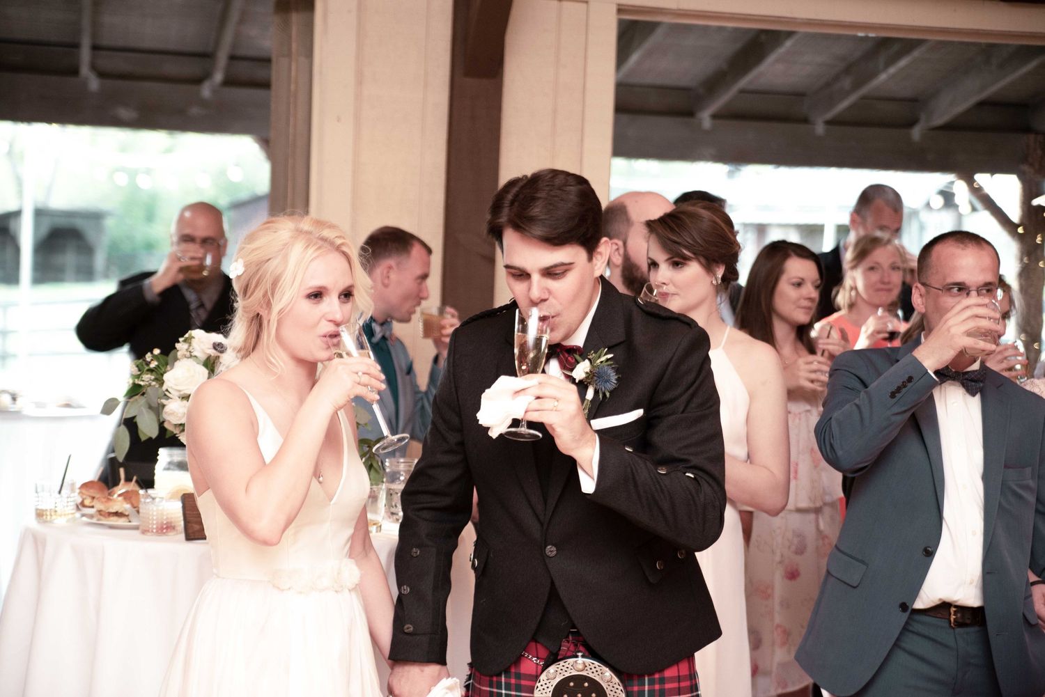 Bride and groom sipping drinks at wedding reception surrounded by guests celebrating in elegant venue.