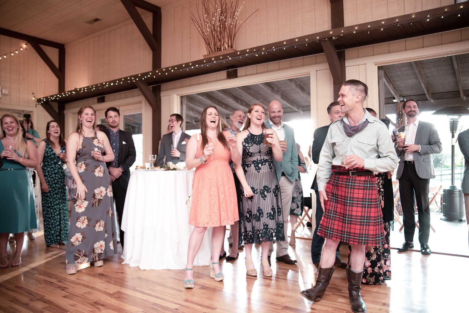 Wedding guests laughing and celebrating in a rustic barn venue, with a man in a red tartan kilt holding a drink.
