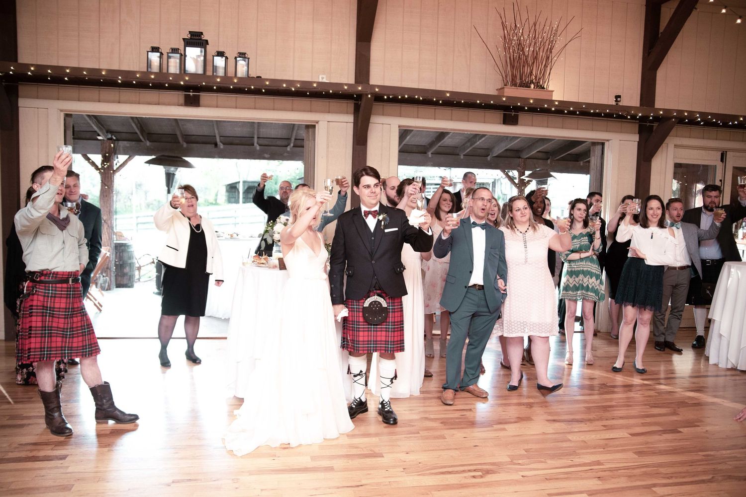 Wedding party celebrating on dance floor, bride in white gown, groom in kilt, guests cheering in rustic barn venue.