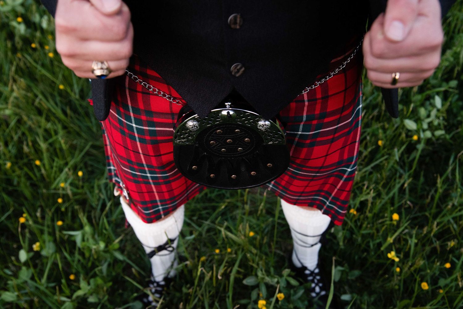 Man wearing traditional Scottish red tartan kilt with sporran and white socks standing in green grass.