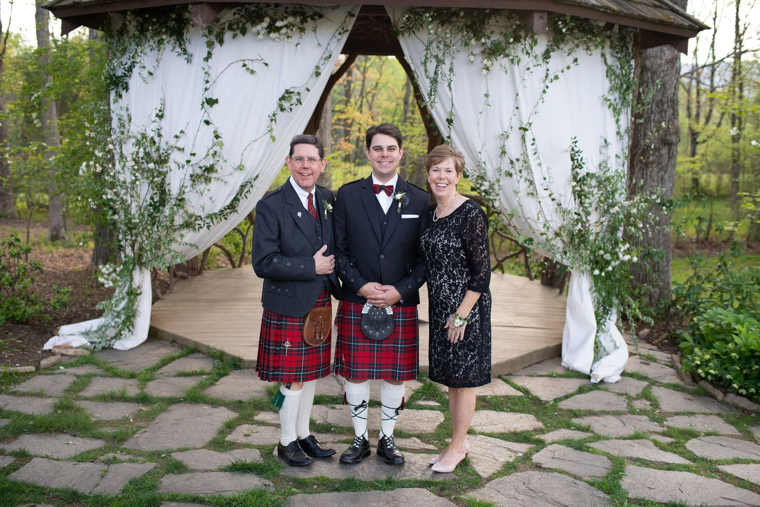 Groom in tuxedo poses with parents, father in red tartan kilt, at outdoor wedding gazebo with white draping.
