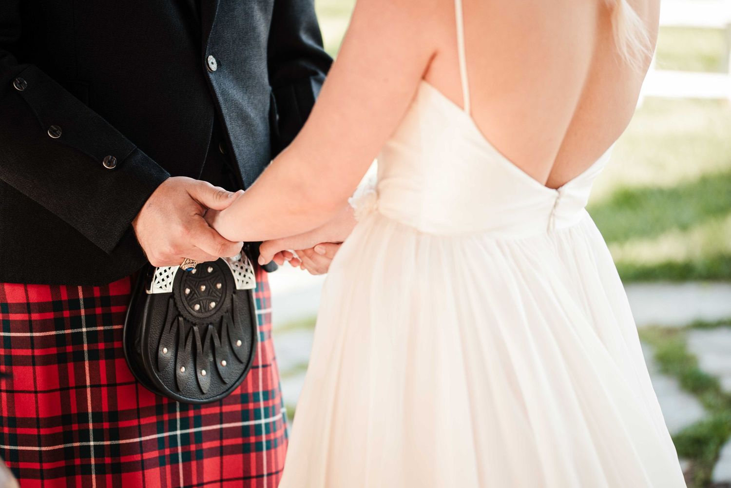 Couple holding hands at wedding, groom in red Royal Stewart tartan kilt with sporran, bride in elegant white backless gown.