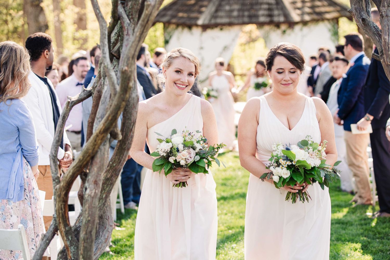 Two smiling bridesmaids in white dresses carrying floral bouquets walk at an outdoor wedding ceremony.