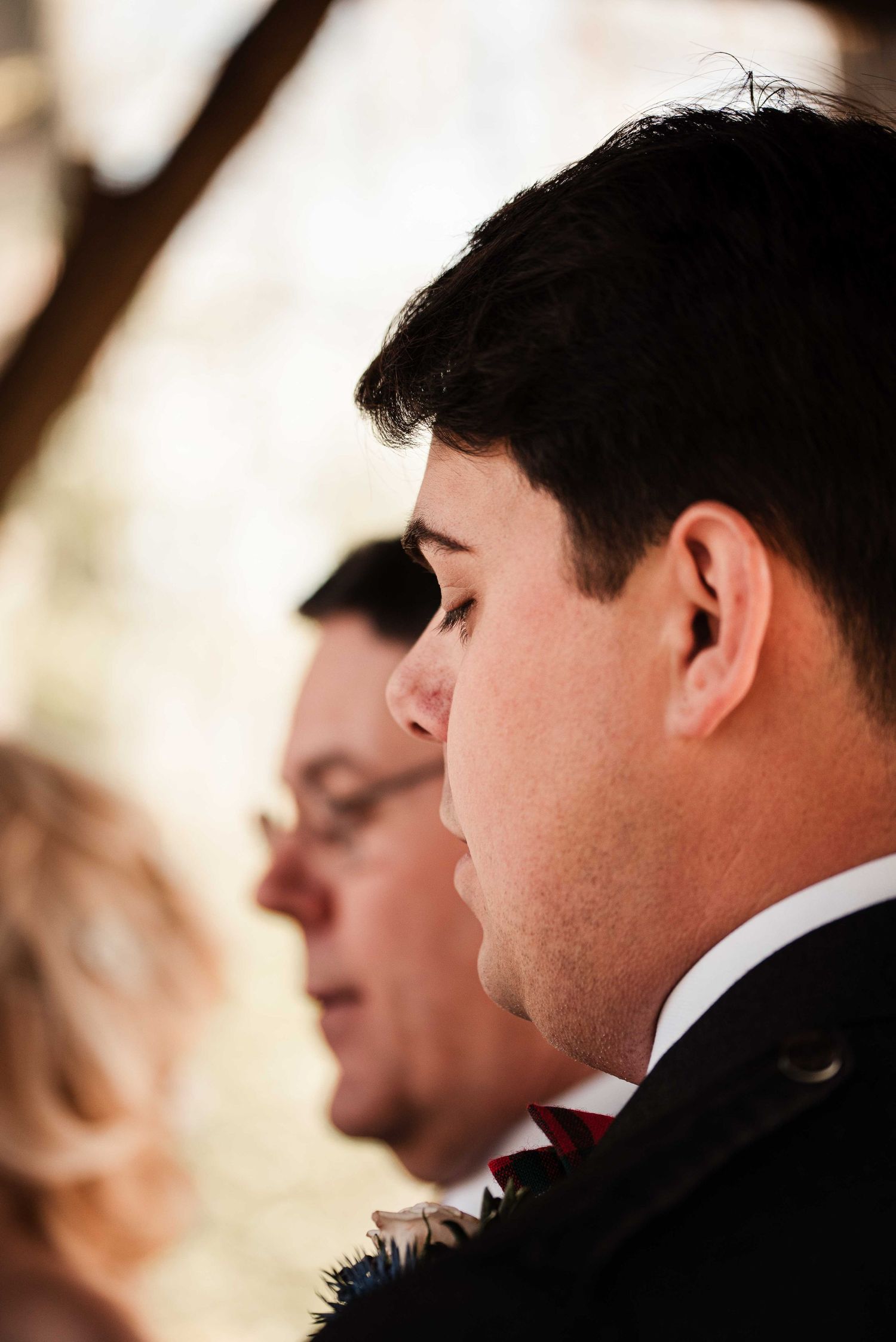 Side profile of a groom in a dark suit with red tie at an outdoor wedding ceremony.