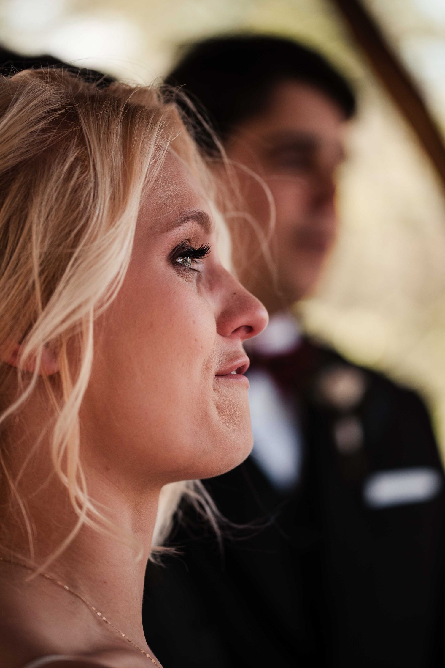 Emotional blonde bride crying tears of joy during wedding ceremony, with groom in dark suit visible in background.