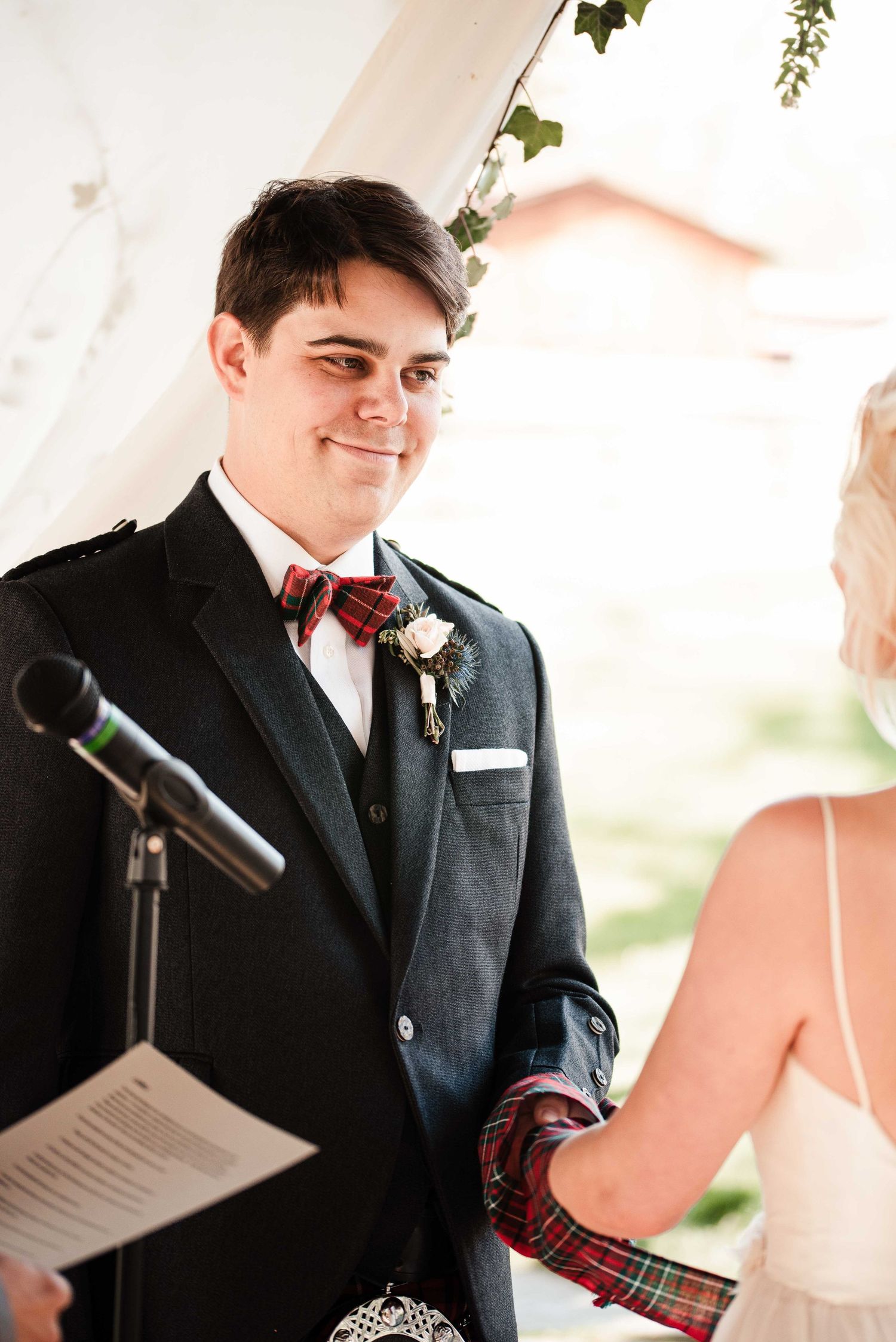 Groom in dark suit with red tartan bow tie smiling at bride during outdoor wedding ceremony.