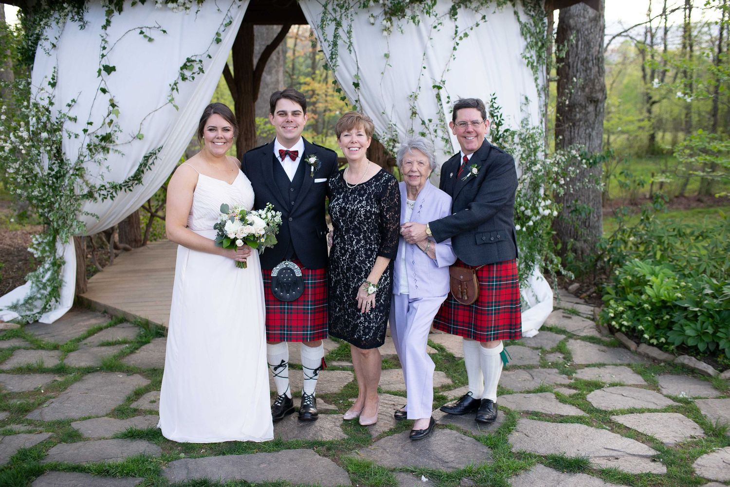 Wedding party poses outdoors with bride in white gown and two men in red kilts under a floral arch on stone path.