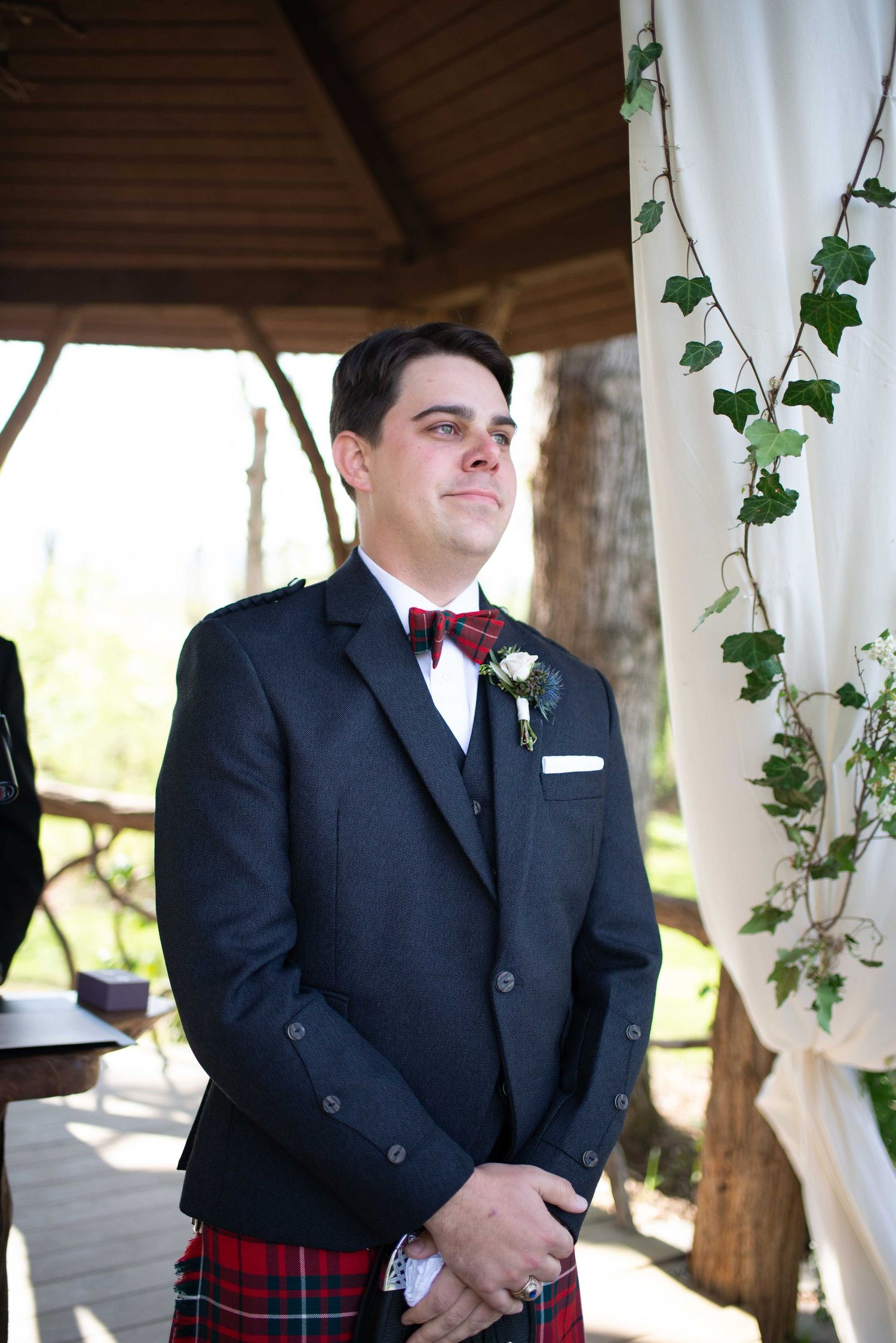 Groom in navy suit, red tartan kilt, and bow tie stands at outdoor wedding altar decorated with ivy.