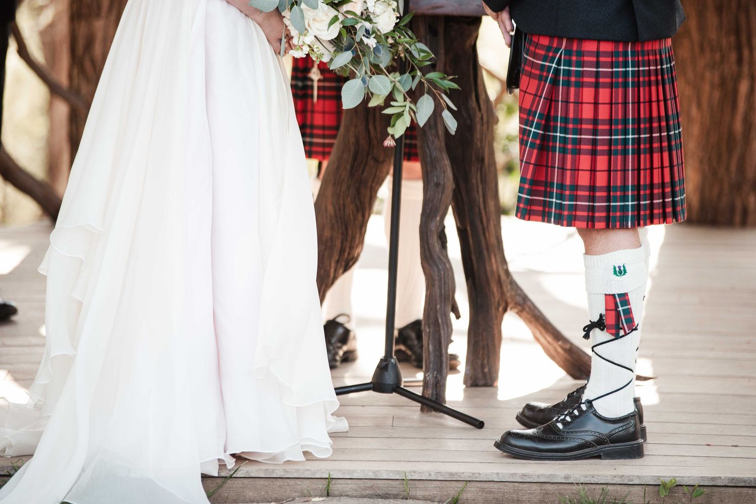 Wedding couple in Scottish tartan kilts and white bridal gown standing on wooden deck with bouquet.