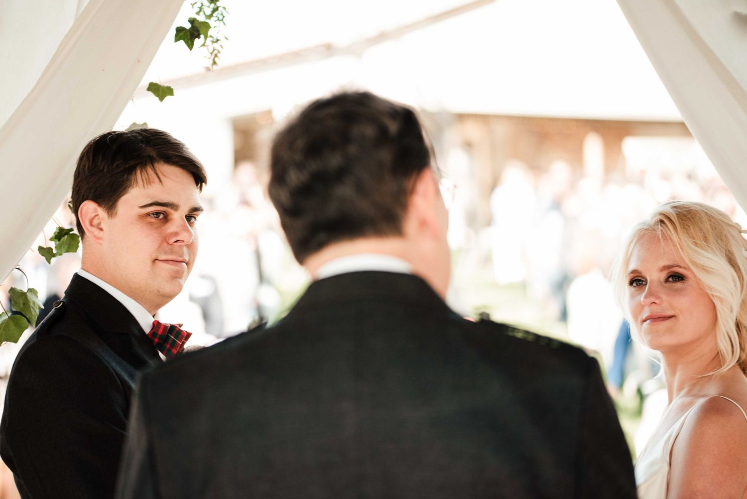 Groom in dark suit smiles at bride in white dress during outdoor wedding ceremony under white draping.