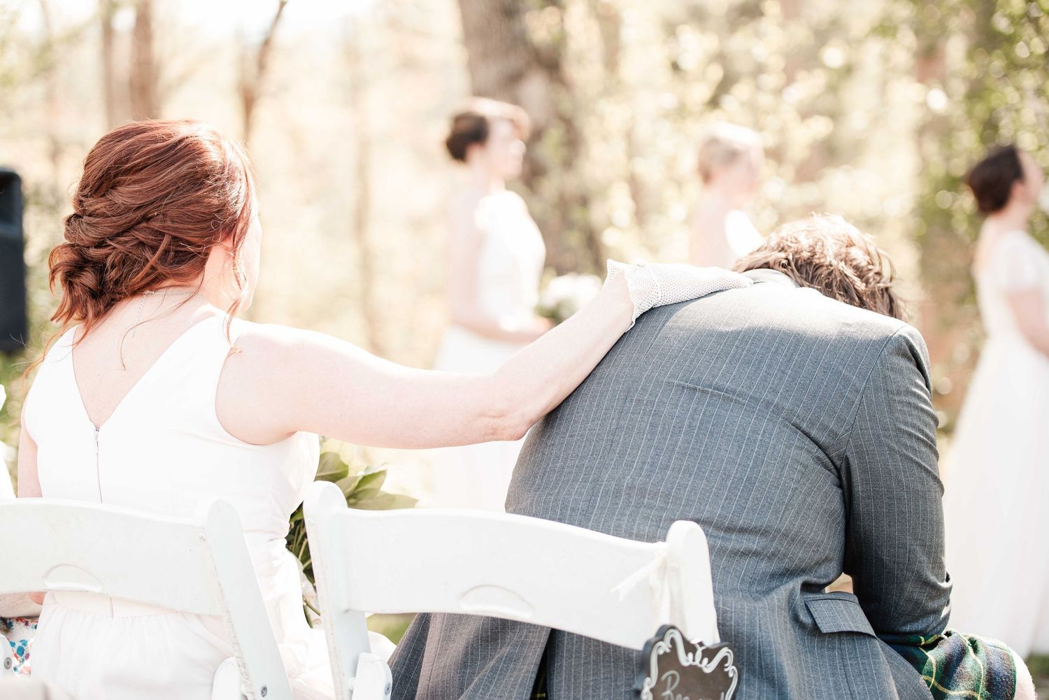 Bride with red updo rests hand on groom's shoulder during outdoor wedding ceremony, seated on white chairs.