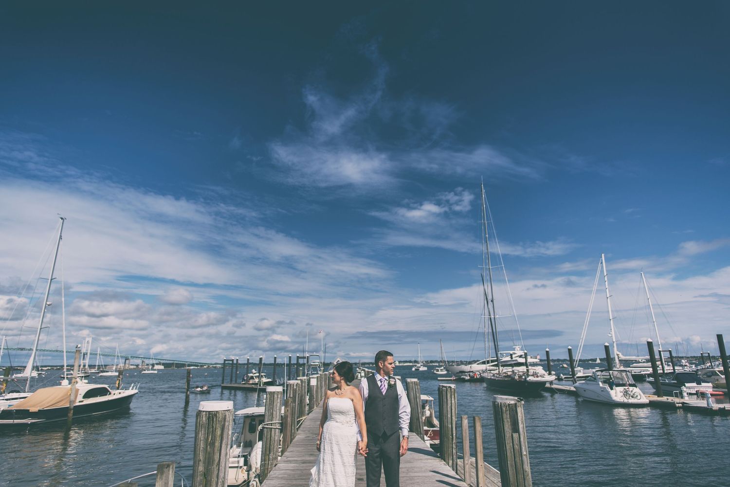 Bride and groom stand on marina dock with sailboats under blue sky.