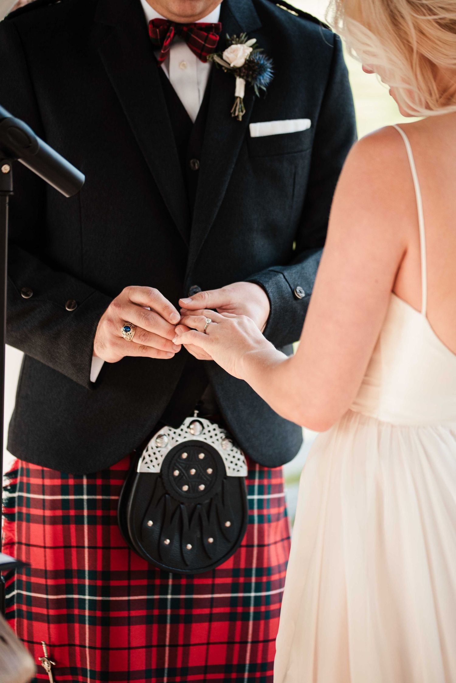 Groom in red tartan kilt and bow tie exchanges rings with bride in white dress during wedding ceremony.