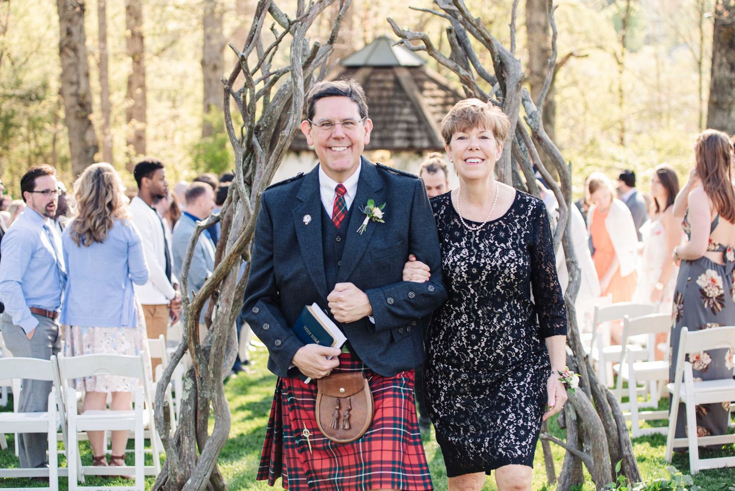 Couple in Scottish kilt and floral dress walking down outdoor wedding aisle surrounded by guests.