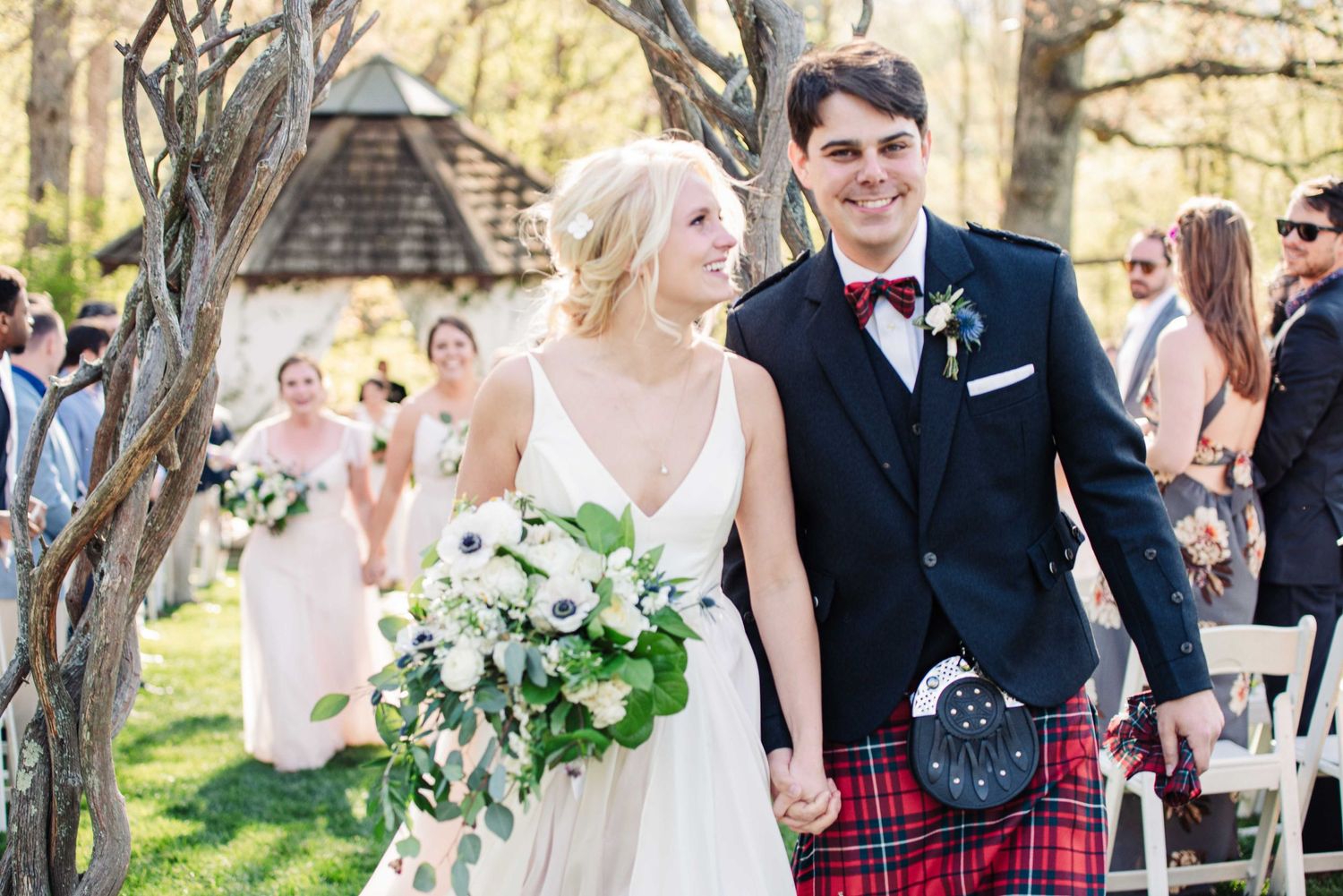 Smiling bride and groom walking down the aisle, groom wearing a tartan kilt, bride holding a white floral bouquet.